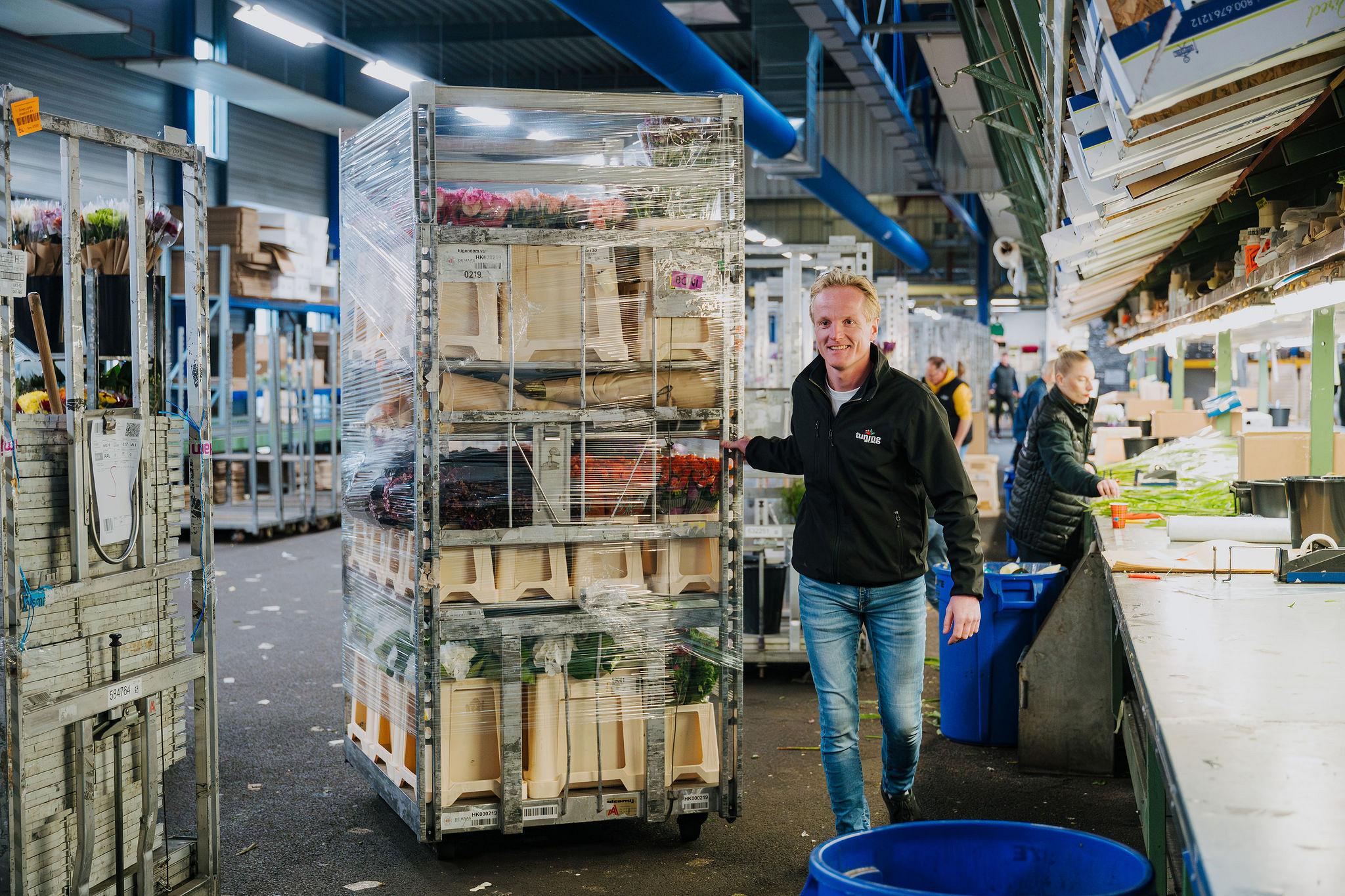 Employee walking through a flower processing facility while pushing a rolling cart with wrapped flower boxes past a conveyor belt where colleagues are assembling bouquets.