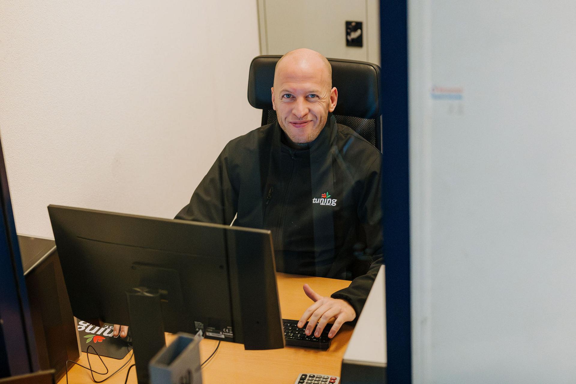 Smiling Tuning BV employee sitting at an office desk behind a glass service window, wearing a black jacket with the company logo.