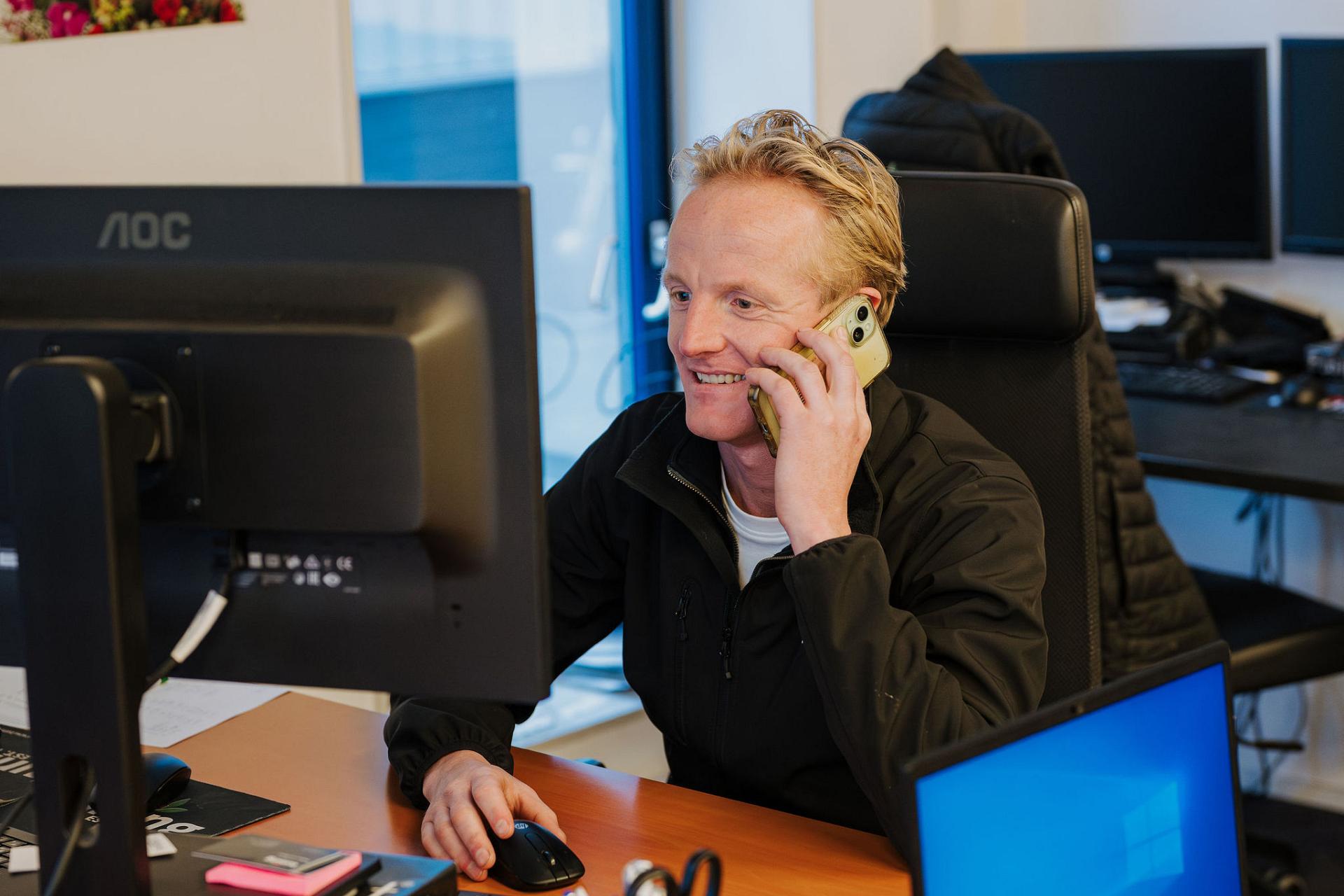 Smiling Tuning BV account manager handling a client call on a mobile phone while working at a desk with multiple screens in a bright office.