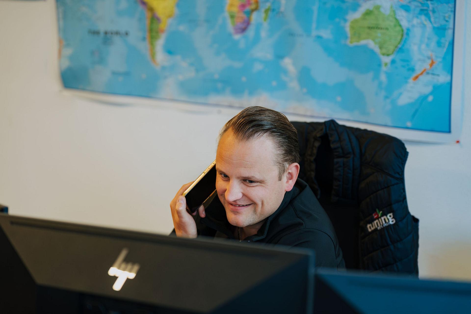 Smiling Tuning BV employee speaking on a mobile phone at an office desk, with a large world map hanging on the wall in the background.