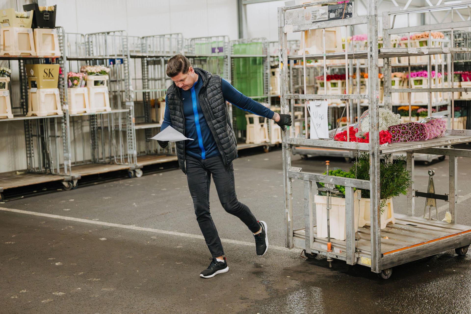 Warehouse employee checking an order list while moving a roll container loaded with fresh flowers in a busy logistics distribution center.
