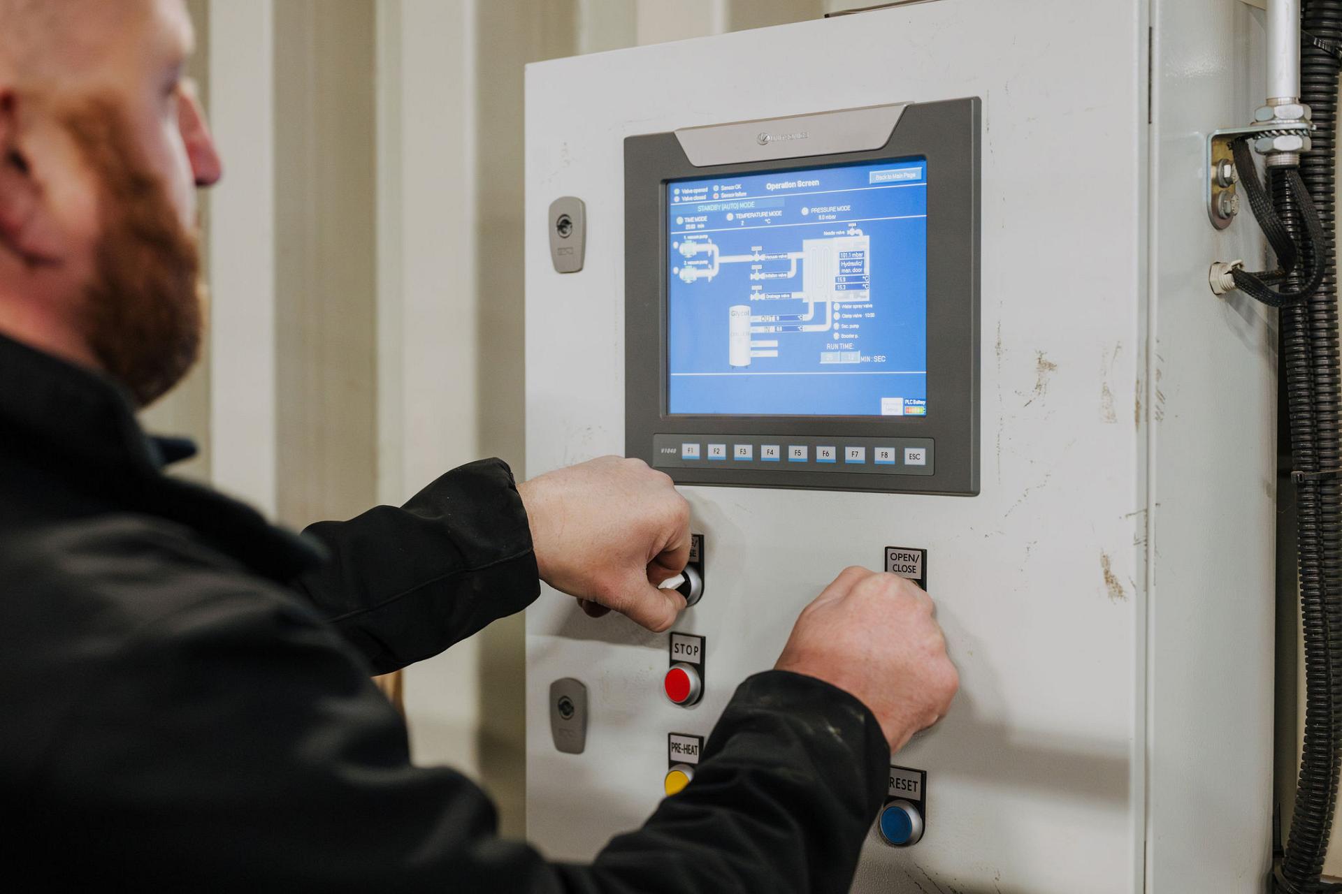 Technician operating an industrial control panel with a digital display showing a schematic operation screen for climate or pressure control.