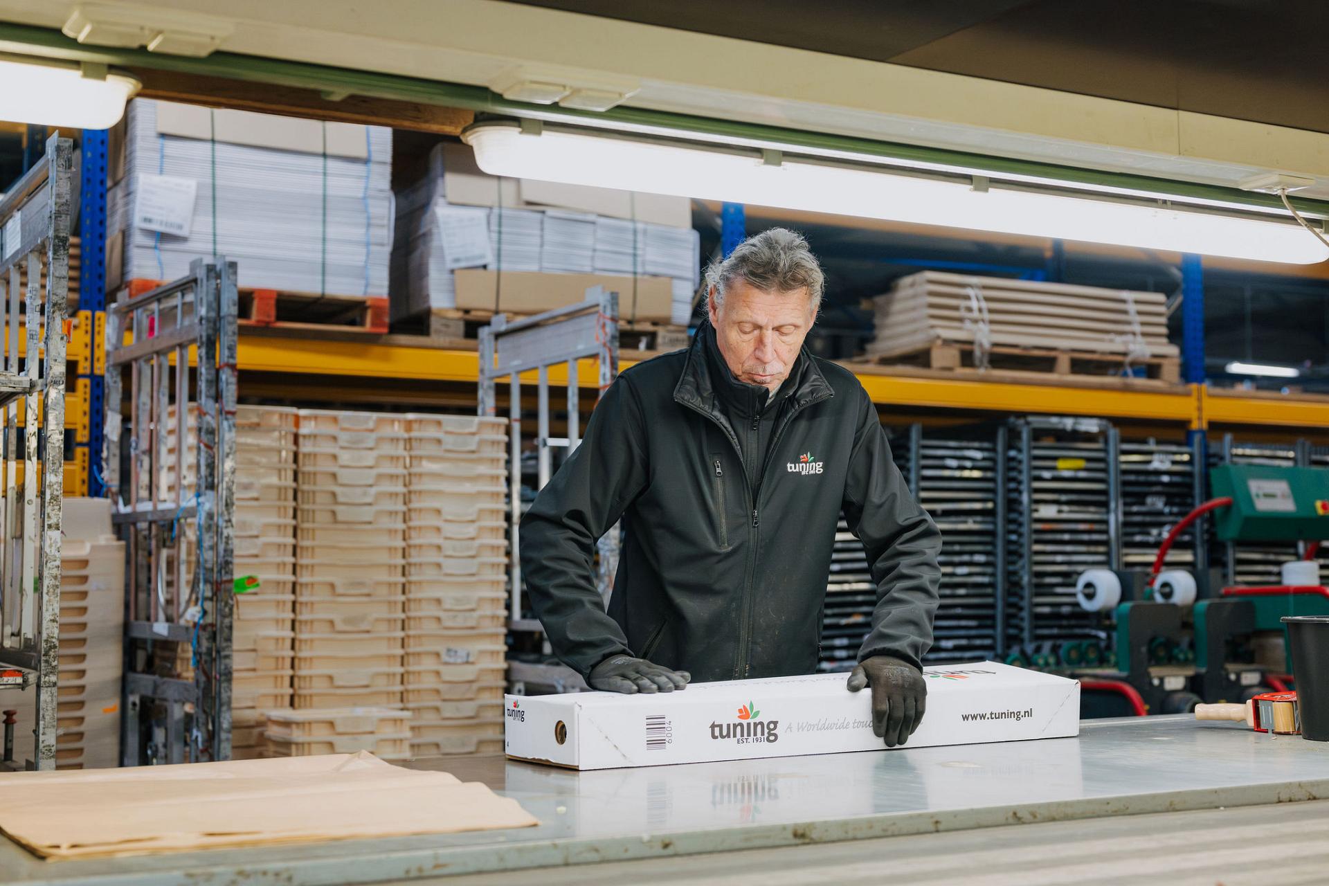 A man in a warehouse works at a packing table while handling a long white shipping box labeled with the Tuning company logo.