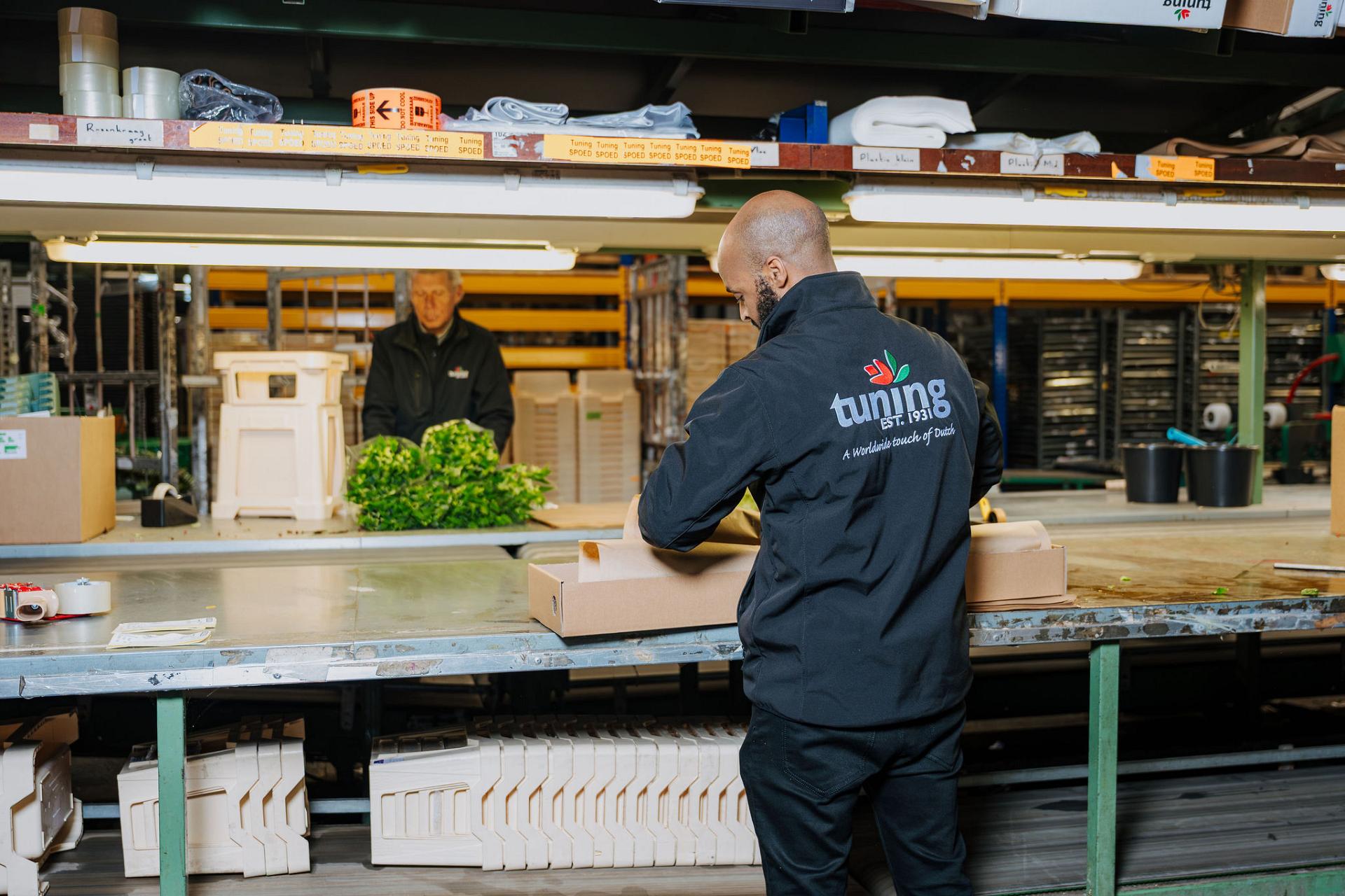 Employee in a flower processing facility packing a cardboard box at a workbench, with another worker and stacked flower crates visible in the background.
