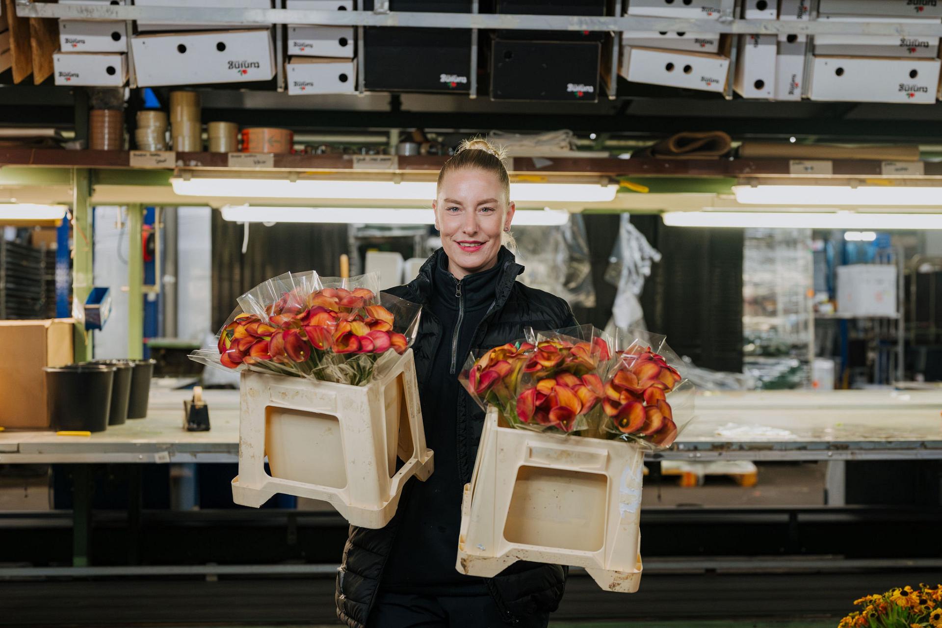 A warehouse employee holds two crates of packaged flowers and smiles in a flower processing workspace.