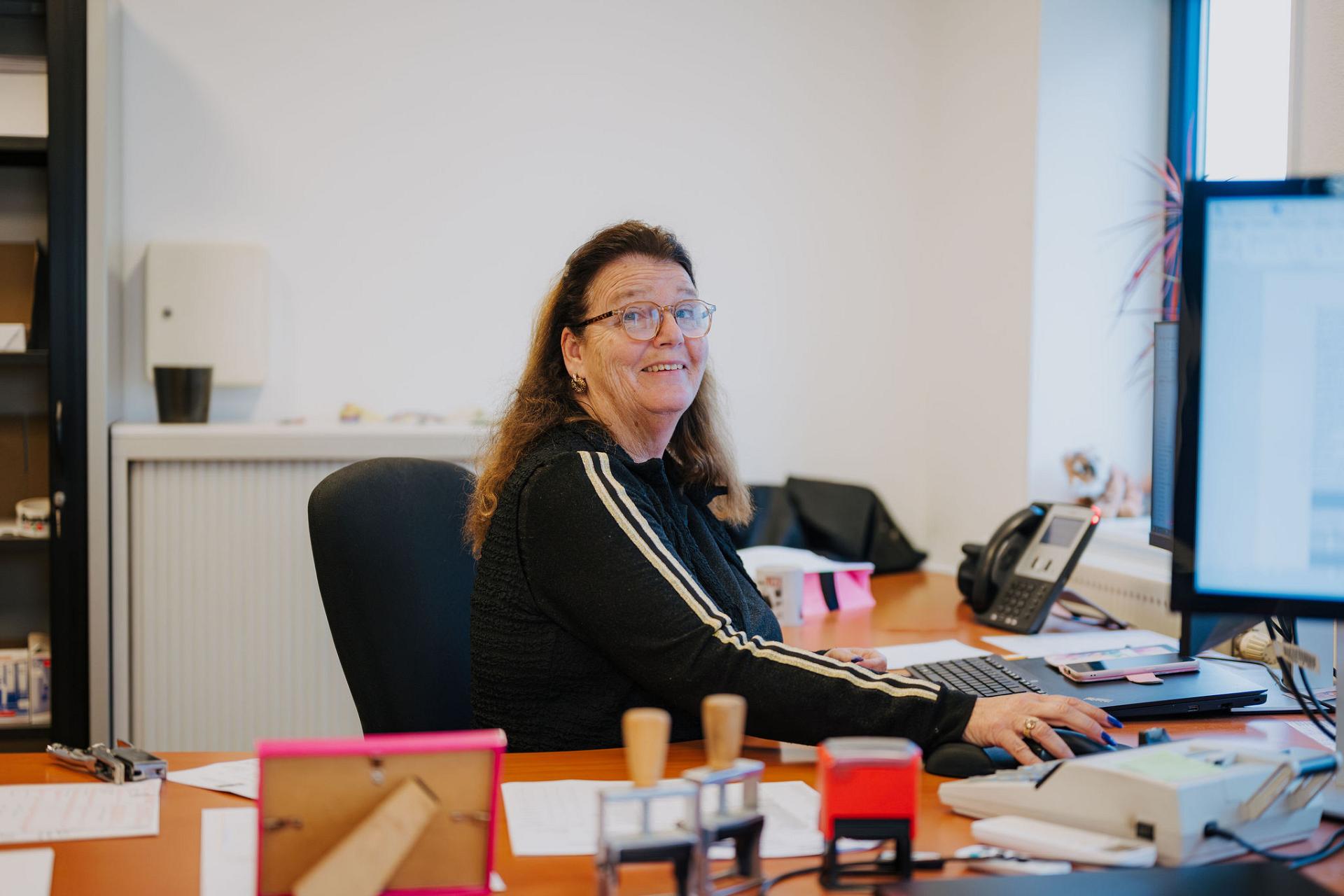 An office employee smiles while working at a desk with a computer, paperwork, and office equipment.