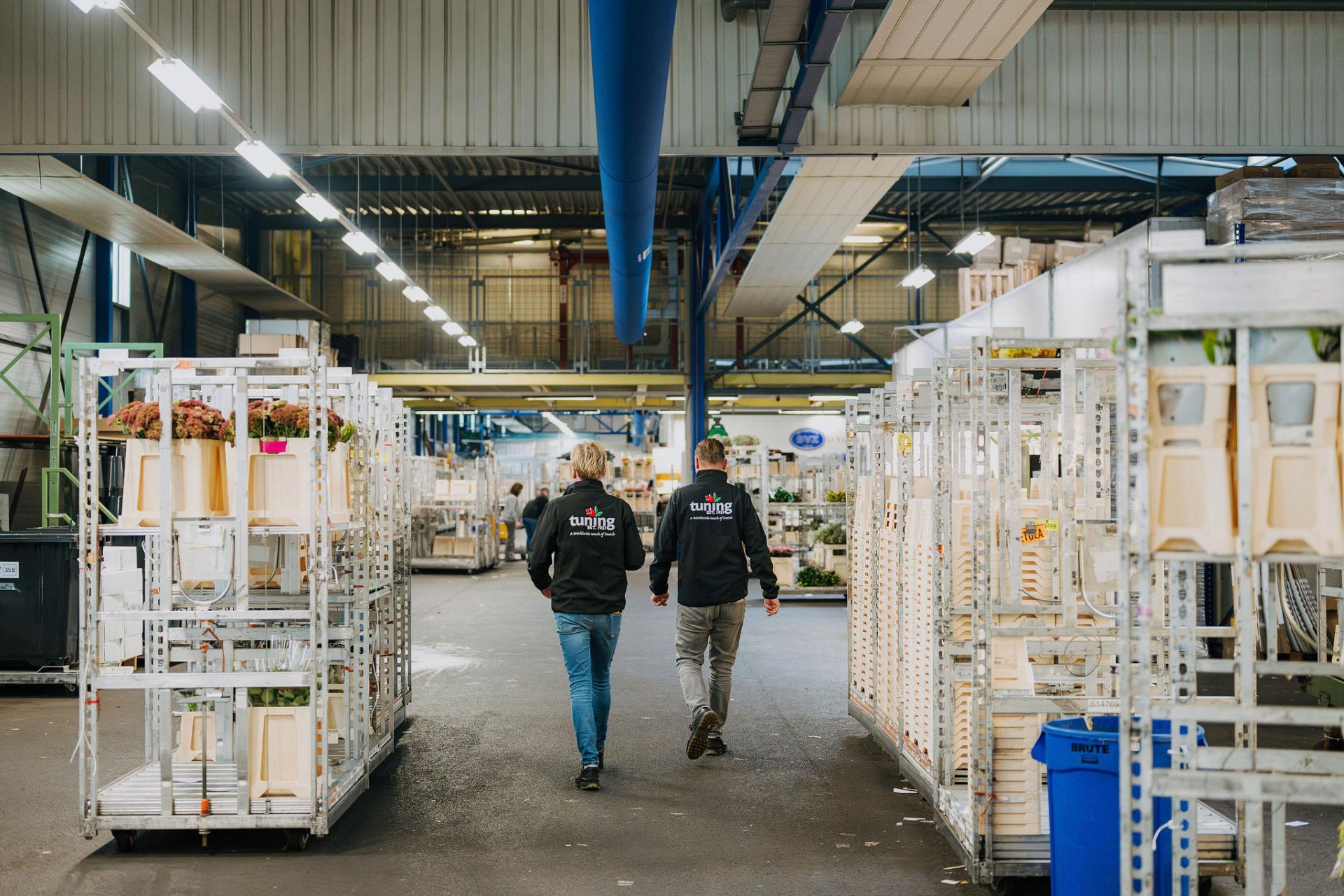 Two warehouse workers walk down a large flower distribution hall lined with rolling racks and crates.