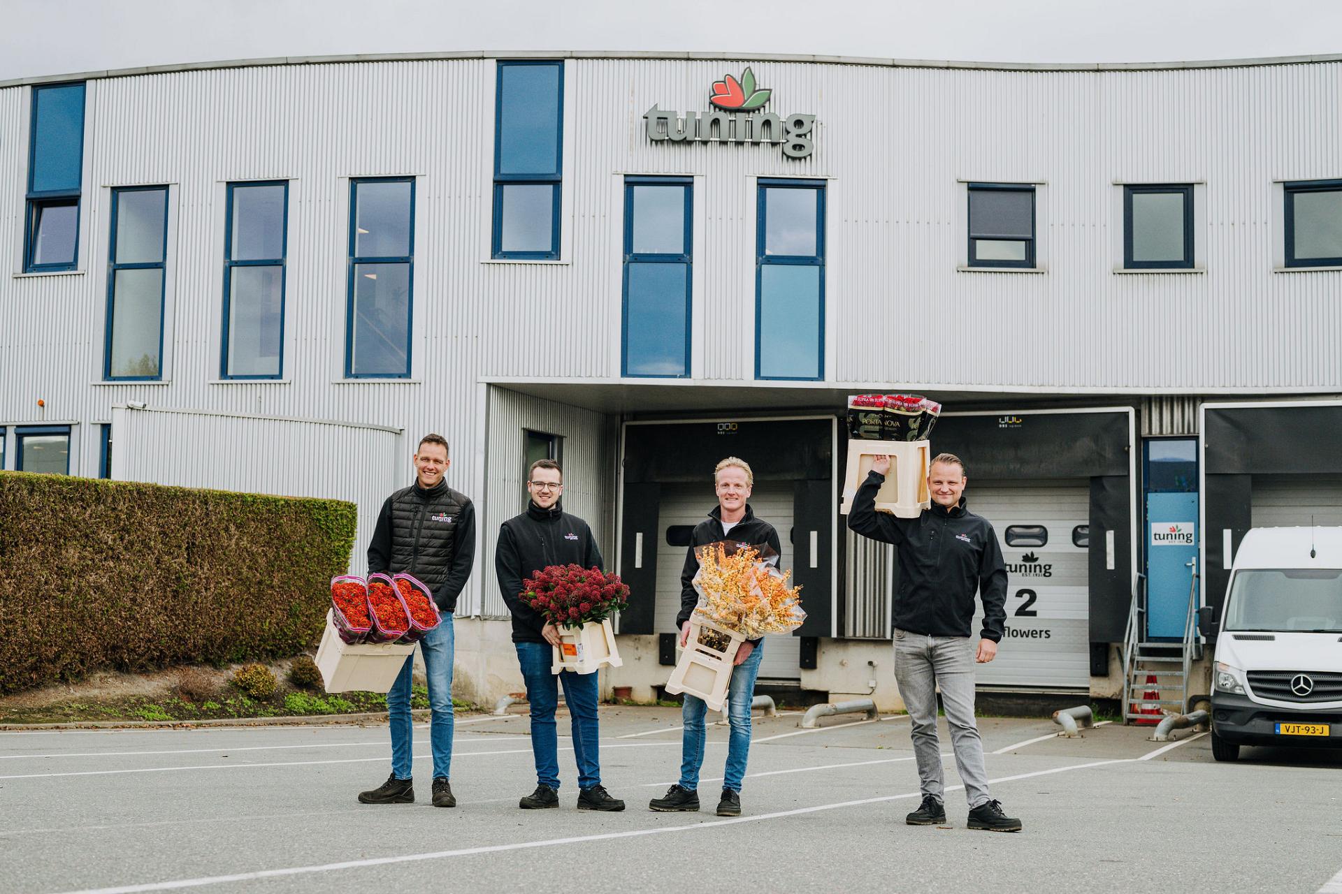 Four employees standing in front of the Tuning company building, holding crates filled with different types of colorful flowers and smiling at the camera.