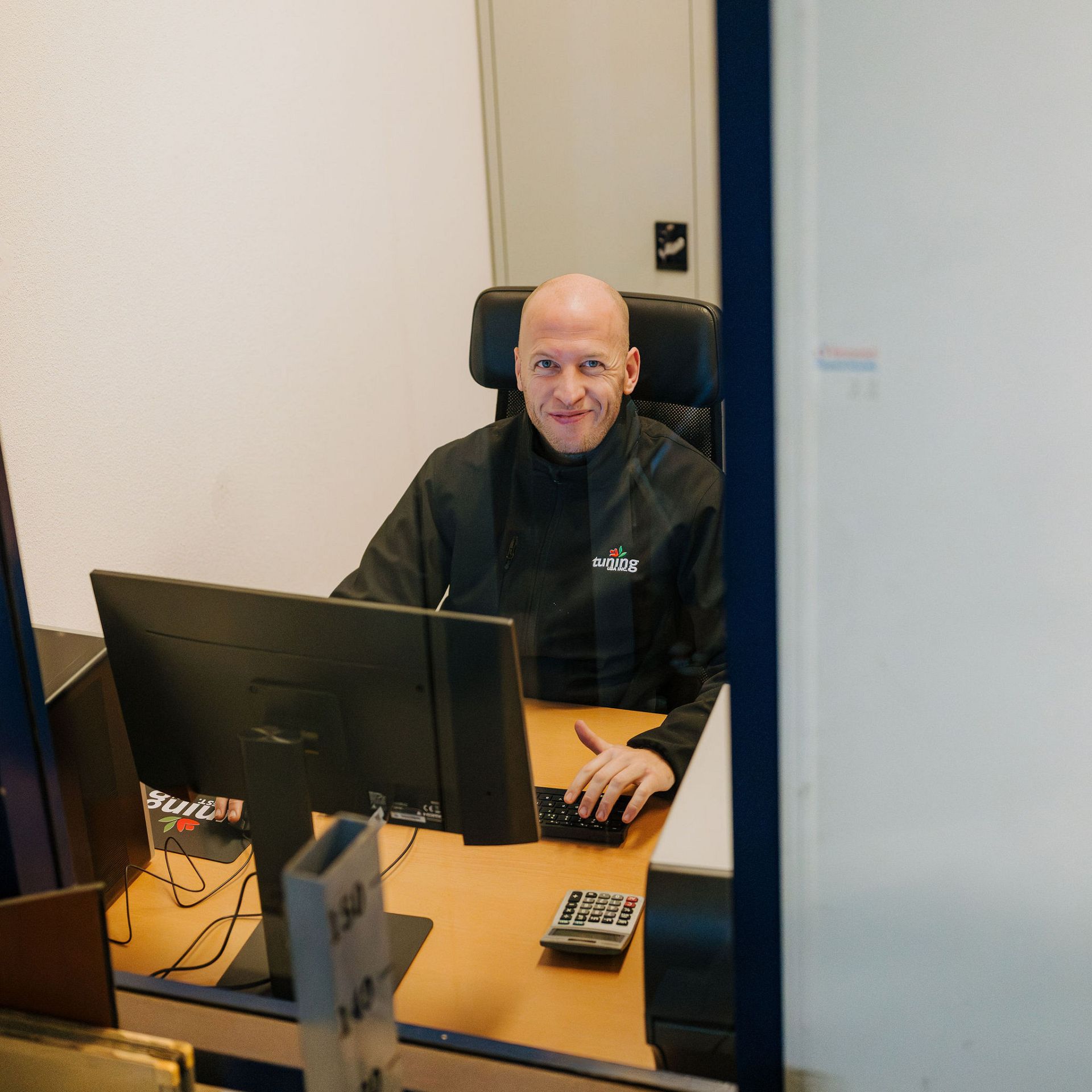 Smiling Tuning BV employee sitting at an office desk behind a glass service window, wearing a black jacket with the company logo.
