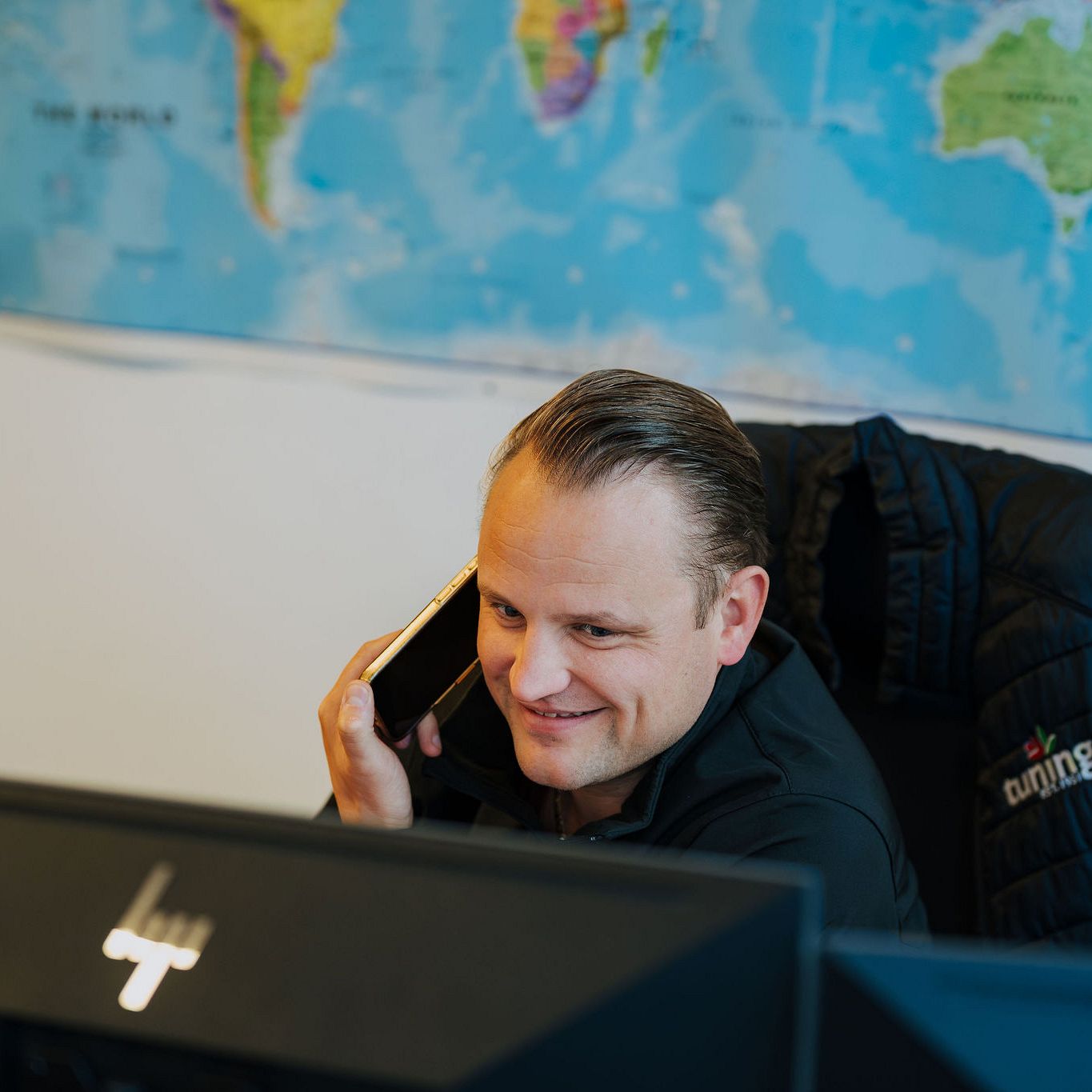 Smiling Tuning BV employee speaking on a mobile phone at an office desk, with a large world map hanging on the wall in the background.
