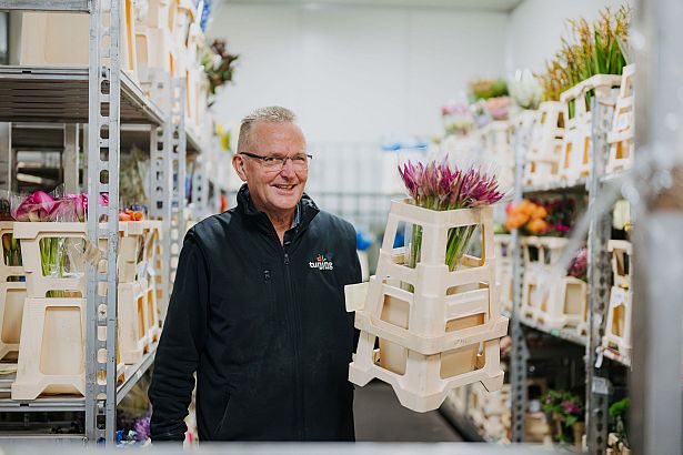 Employee in a flower processing facility holding two crates of wrapped orange flowers and smiling at the camera.